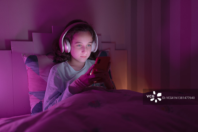 年轻女孩坐在床上使用手机和无线耳机。图片素材
