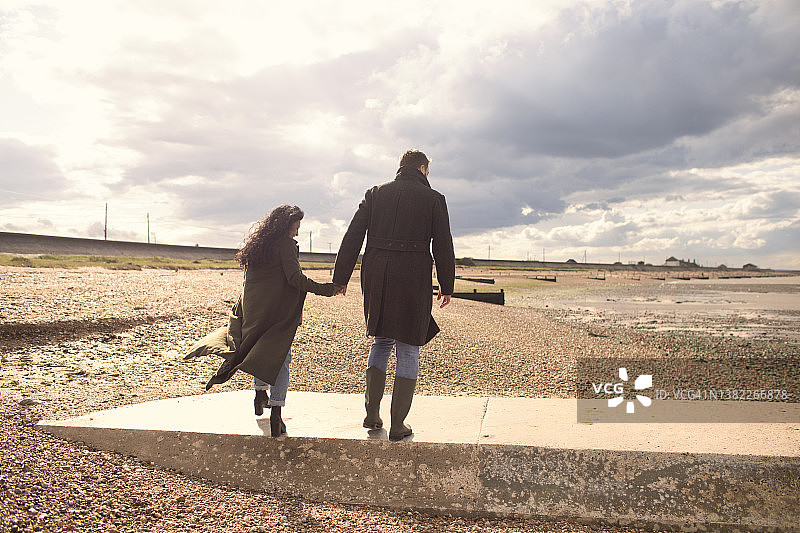 穿着冬衣的情侣手牵着手走在阳光明媚的海滩上图片素材