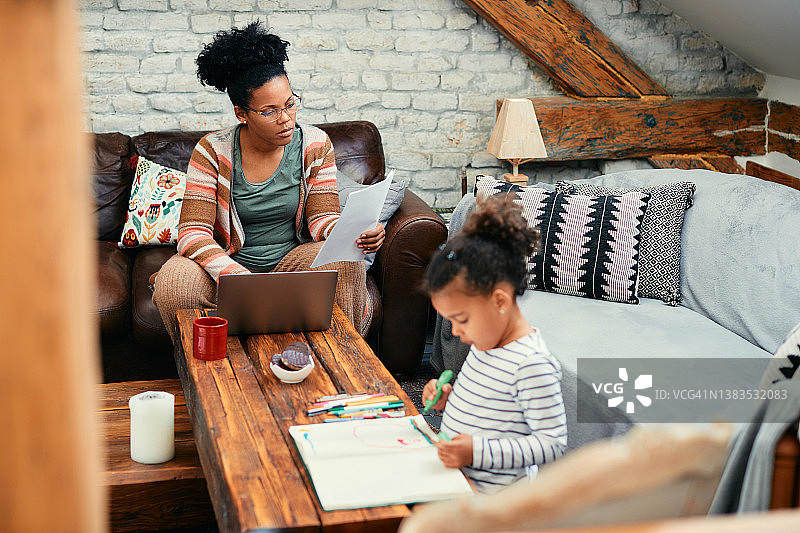 黑人职业母亲分析文件和使用笔记本电脑，而她的女儿在家里在纸上涂色。图片素材