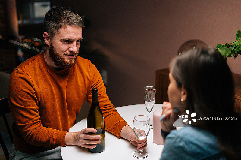 在节日餐桌上，年轻男子与妻子交谈时将酒倒入酒杯的高视角镜头。图片素材