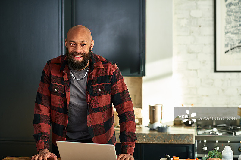 一名留着胡子的中年黑人男子在厨房里用笔记本电脑，面带微笑图片素材