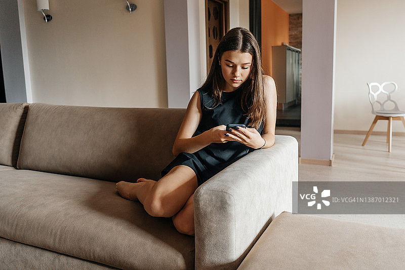 一个拿着手机的无聊少女坐在客厅的沙发上。图片素材