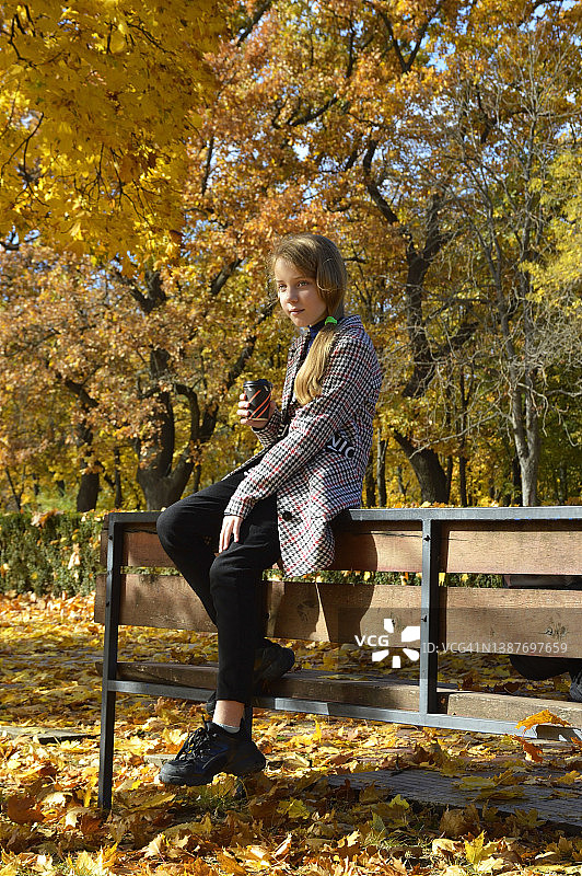 一个女孩穿着格子呢外套，端着一杯咖啡，坐在秋天公园的长椅上图片素材