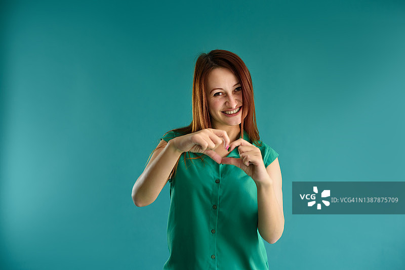 迷人的快乐的红发年轻女子的肖像显示蓝色背景上的心形标志。图片素材