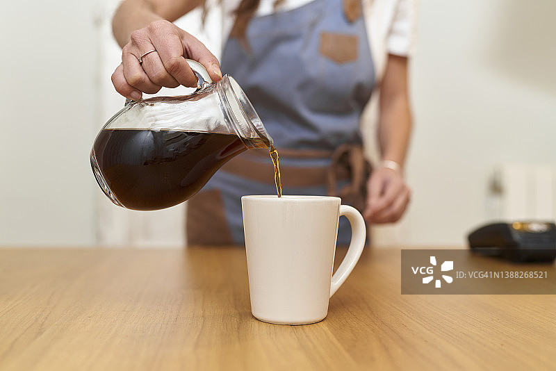 半截特写，一个不认识的女人穿着围裙，站在她家的厨房里，在模糊的背景下把黑咖啡倒进一个白色的咖啡杯图片素材