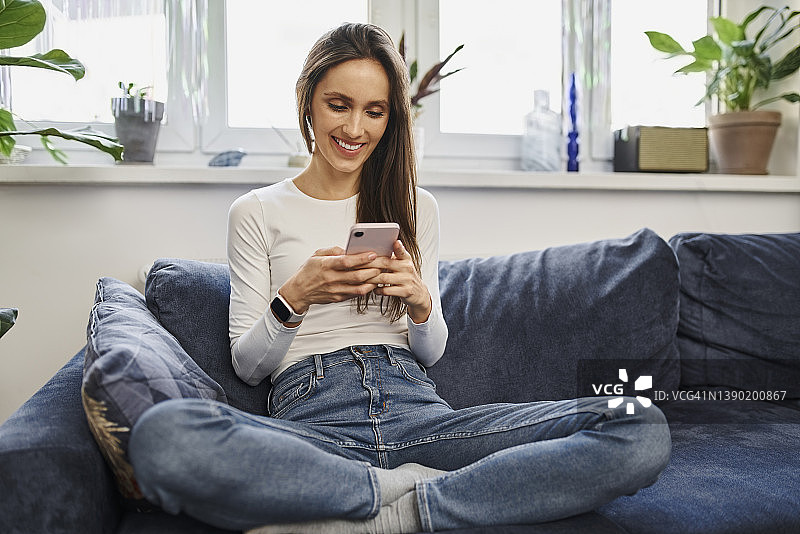微笑的女人用手机盘腿坐在家里的沙发上图片素材