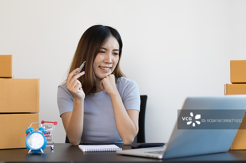 一位年轻的亚洲女性开始了一个小型的电子商务企业，她正在用笔记本电脑工作，并通过互联网与客户聊天。图片素材
