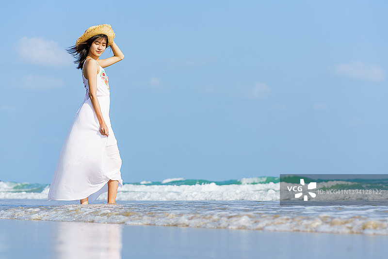 年轻女子在白色的太阳裙和手与帽子在夏天独自走在沙滩上的特写图片素材