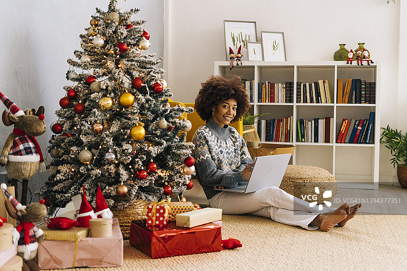 快乐的年轻女子带着笔记本电脑坐在家里的圣诞树旁图片素材