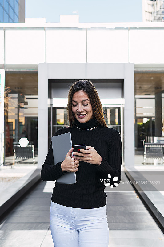 微笑的年轻女商人与笔记本电脑使用智能手机在咖啡馆前图片素材