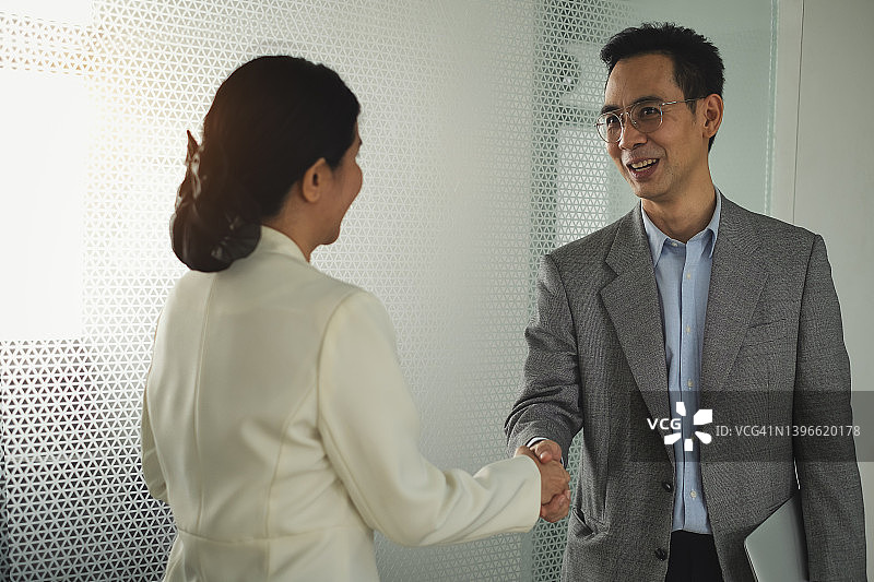微笑的商业伙伴男人在聪明的办公室西装握手与商业女人，而持有笔记本电脑前不清楚的镜子墙的会议室在办公室图片素材