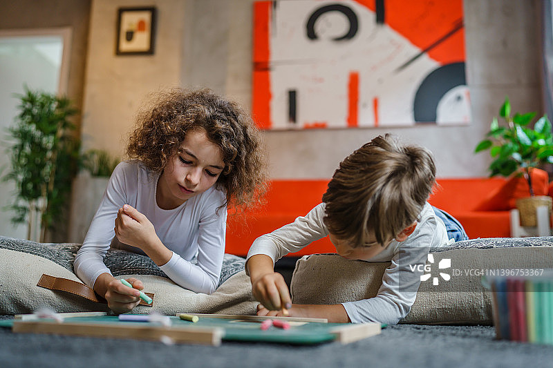 小男孩和小女孩在家里的绿板上画画，两个孩子兄弟姐妹或朋友躺在地板上用粉笔休闲教育真人家庭概念拷贝空间图片素材