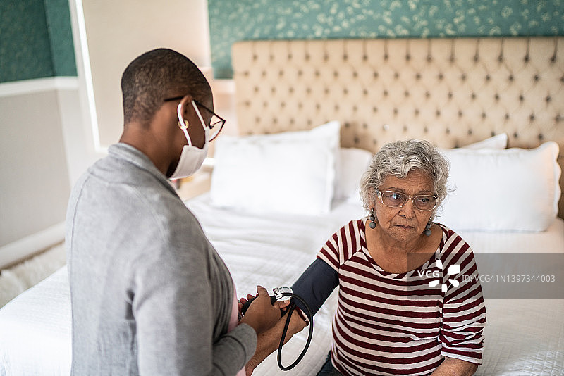 医生在卧室里测量一位老年妇女的血压——戴着口罩图片素材