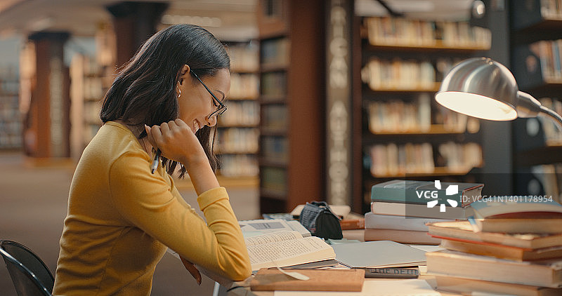 快乐的学生独自在图书馆看书。年轻的女士微笑着，享受着她的学习。混血女性在做一个项目图片素材