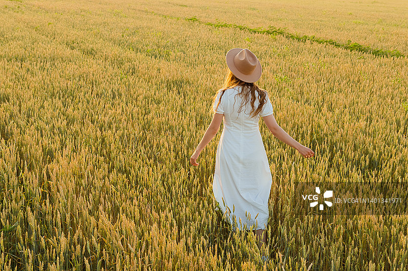 年轻美丽的千禧一代女子身穿白色长裙，帽子一顶，站在夕阳下金色的麦田里，眺望远方。图片素材