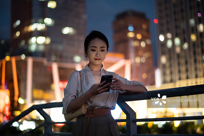 亚洲年轻漂亮的女人晚上在城市街道上使用智能手机图片素材
