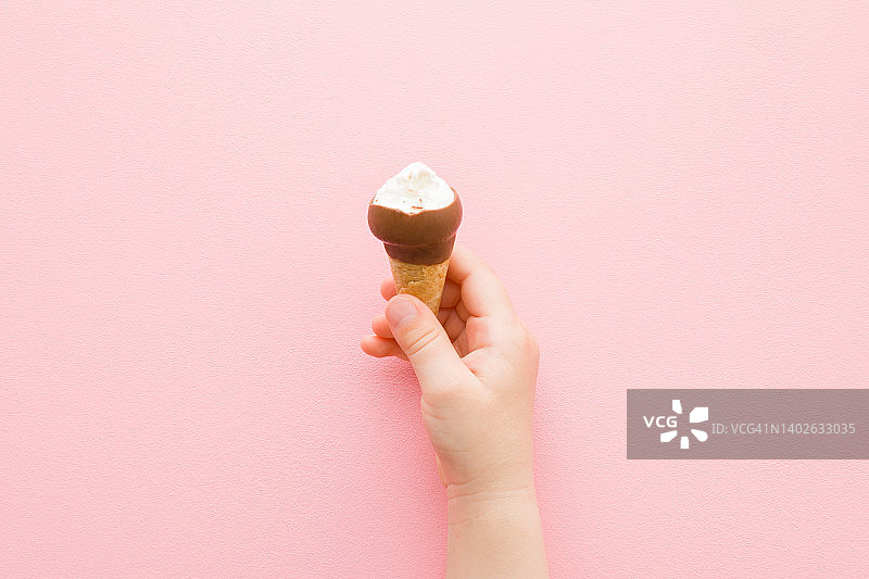 女婴手握华夫饼筒白色香草冰淇淋与黑巧克力釉在浅粉红色的桌子背景。柔和的颜色。特写镜头。冷甜的零食。咬的食物。自顶向下的观点。图片素材