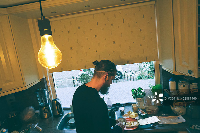 一个留着长卷发的男人正站在厨房的窗边忙着做饭。休息日在家吃早餐图片素材