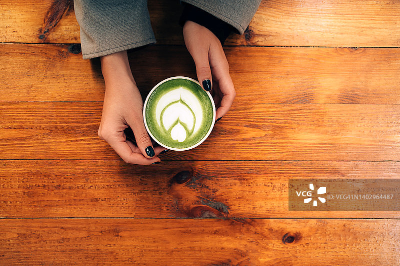 女人的手握陶瓷杯抹茶绿茶拿铁与泡沫艺术在木桌上。您的设计背景与复制空间。摄影从上面图片素材