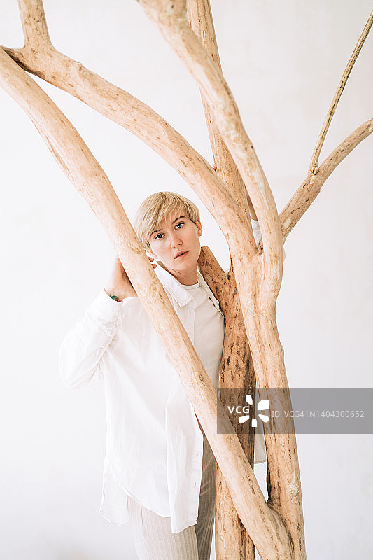 美丽的真实的女人与短的金发拥抱树在室内。她穿着一件白衬衫。自然美容护肤理念图片素材