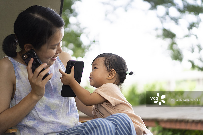 坦诚积极的情绪亚洲妈妈和女儿在户外的大树下和复印空间使用电话图片素材