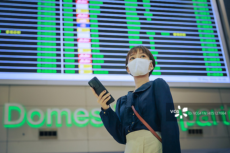 年轻的亚洲女商人戴着口罩手持手机站在机场出境区。商务旅行的概念。图片素材