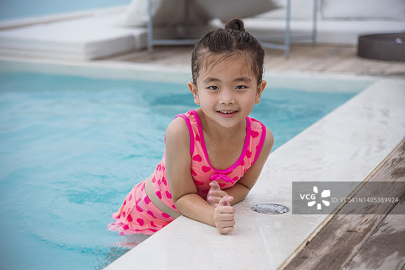 穿着粉色泳衣的少女肖像在游泳池嬉戏夏日时光用粉色泳衣，蓝色的水在酒店游泳池室外。蹒跚学步的女孩孩子笑笑看着镜头。快乐女孩游泳俏皮图片素材