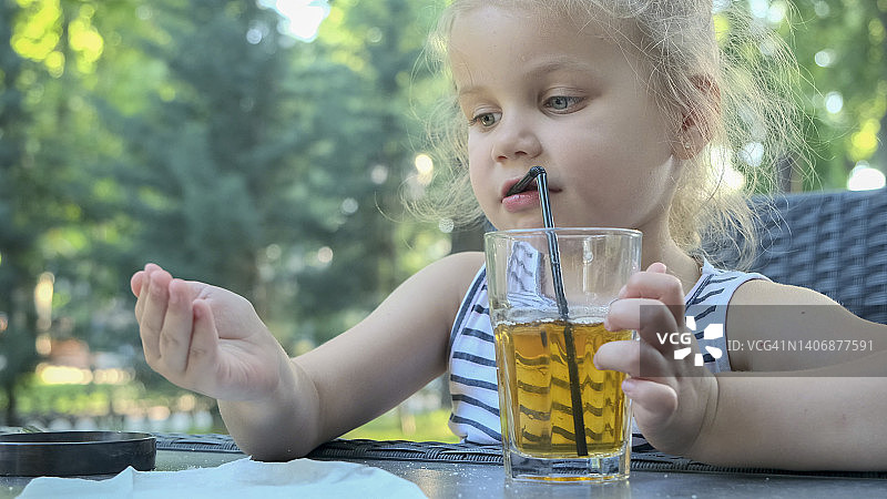 可爱的小女孩用吸管喝果汁。特写肖像的金发女孩喝果汁从玻璃通过鸡尾酒吸管坐在街头咖啡馆的公园。图片素材