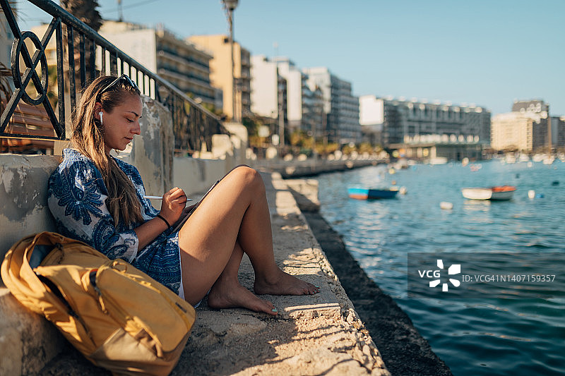 一位戴着无线耳机的女士坐在海边的一个码头上，用着一台数字平板电脑图片素材