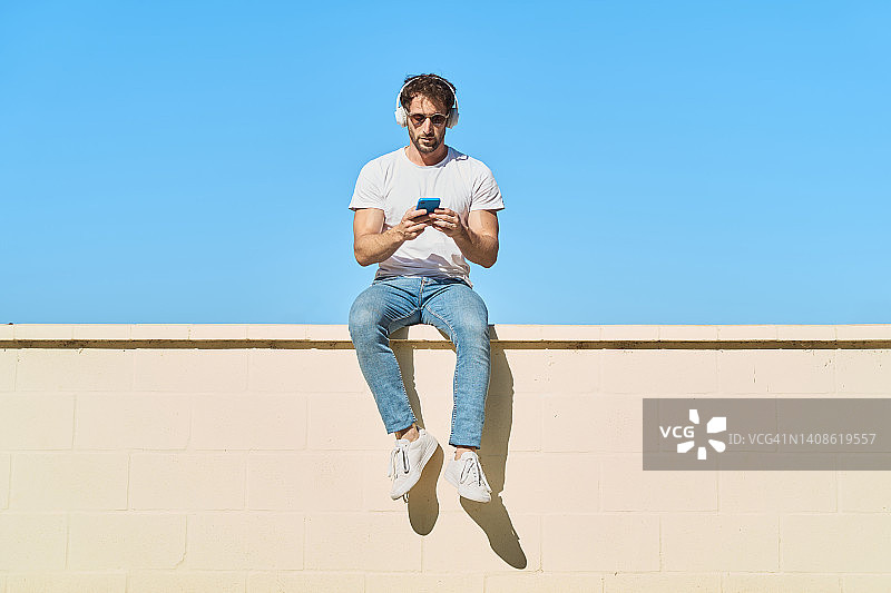 年轻人穿着休闲的衣服，坐在以蓝天为背景的黄色砖墙上，一边用手机一边戴着无线耳机听音乐。图片素材
