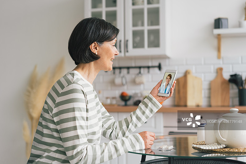 微笑的高级女性在厨房用智能手机与女医生进行在线咨询。远程医疗的概念。大流行期间在家远程办公。寻求帮助和抱怨症状，水平图片素材
