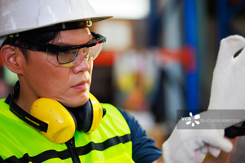 肖像英俊的亚洲男性仓库专业工人在仓库工厂工业安全背心和安全帽头盔。产品在存储的货架上的纸箱。图片素材
