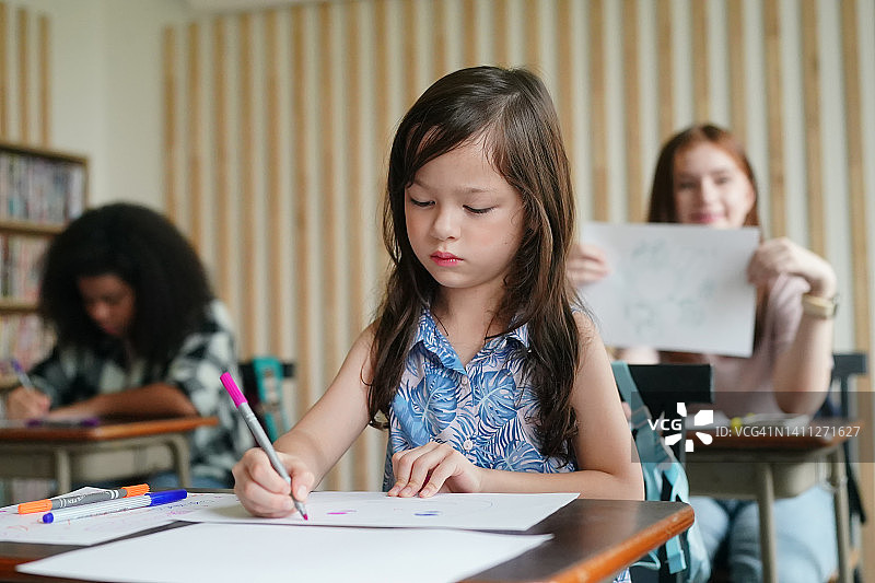 学前班女孩孩子在教室的桌子上用彩色铅笔在白纸上画画图片素材