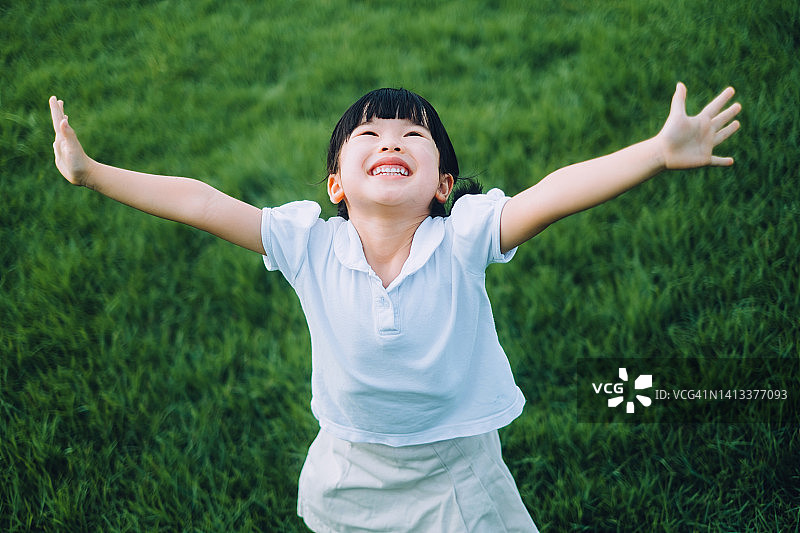快乐的亚洲小女生在公园的草地上玩得很开心。她高兴地笑着，张开双臂。在夏日的户外享受大自然和自由时光。童年的生活方式。无忧无虑，自由和俏皮的概念图片素材