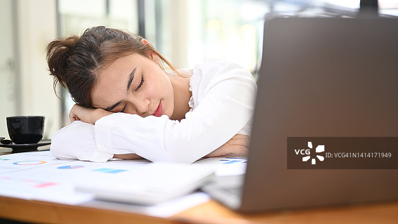 疲惫的女办公室职员在她的办公桌上打盹，她的头躺在她的胳膊上，因为工作累了。图片素材