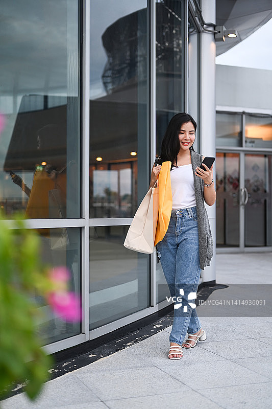 全长肖像的年轻女子与购物袋使用智能手机，走在城市街道在晚上。图片素材
