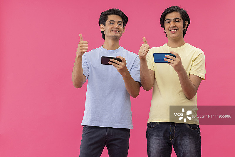 两个十几岁的男孩站在粉色背景下玩智能手机游戏图片素材
