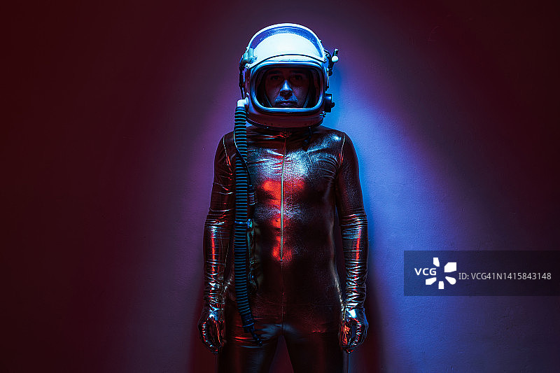 宇航员穿着金色的连体衣和头盔，在蓝色和红色灯光的照射下，看着摄像机。探索的概念，空间，行星，陌生和陌生。图片素材