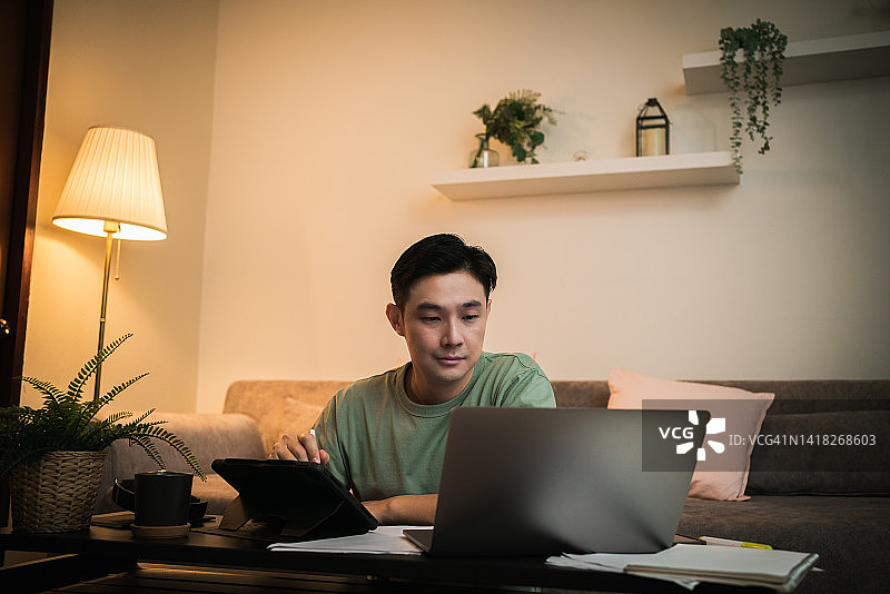 亚洲成年学生在线学习在笔记本电脑上上课，在家里客厅的沙发上喝热咖啡。图片素材