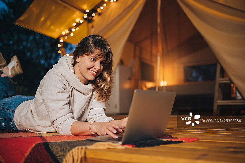 快乐的女人自由职业者使用笔记本电脑在一个舒适的野营帐篷在一个夏天的晚上。户外度假度假的豪华露营帐篷。生活方式的概念图片素材