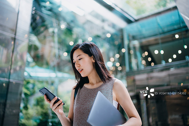 自信和成功的年轻亚洲女商人带着笔记本电脑，同时使用智能手机在路上，通勤上班在市中心区与城市的现代企业建筑。职场中的女性领导力。忙碌的生意图片素材