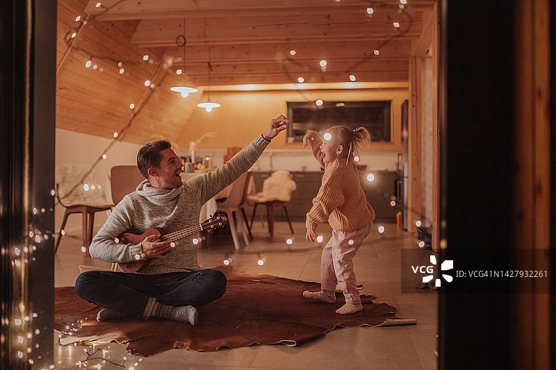 年轻快乐的爸爸坐在客厅里弹吉他，他的小女儿在跳舞。图片素材