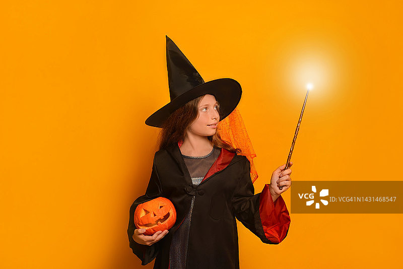 小女孩在巫师学生服装拿着发光的魔术棒或发光魔术棒雕刻万圣节南瓜或杰克o南瓜灯的黄色背景。图片素材