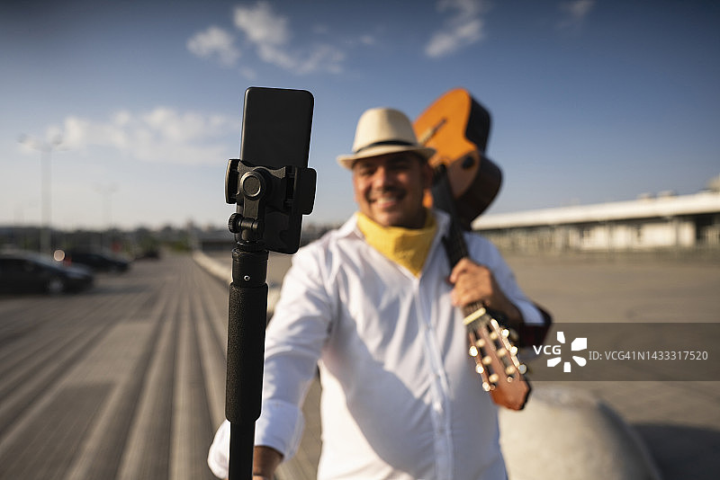 微笑的吉他手拿着吉他在三脚架上通过手机自拍图片素材