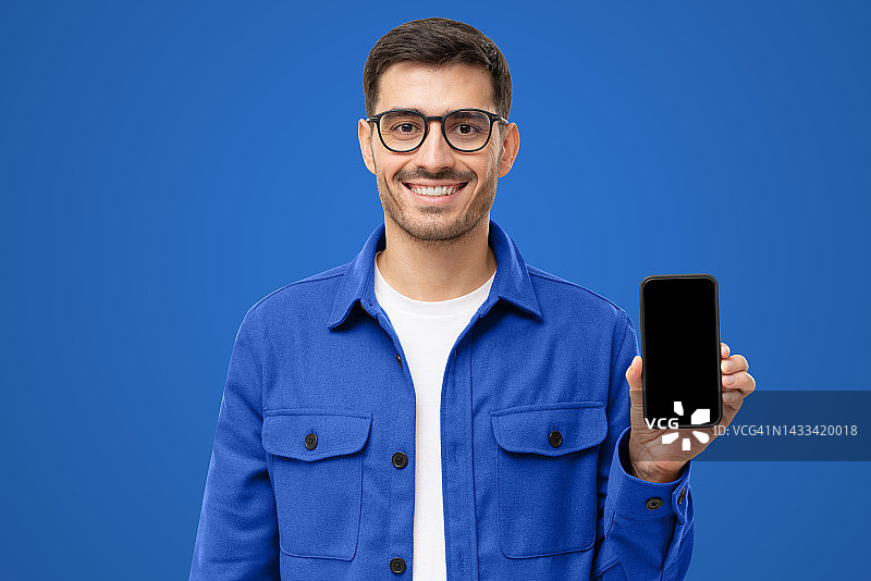 年轻的微笑的男人在休闲衬衫显示空白屏幕的智能手机在手中，孤立的蓝色图片素材