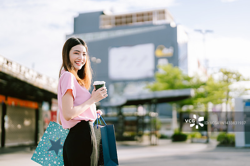 一位年轻的亚洲妇女提着购物袋和咖啡在购物中心前散步，玩得很开心。图片素材