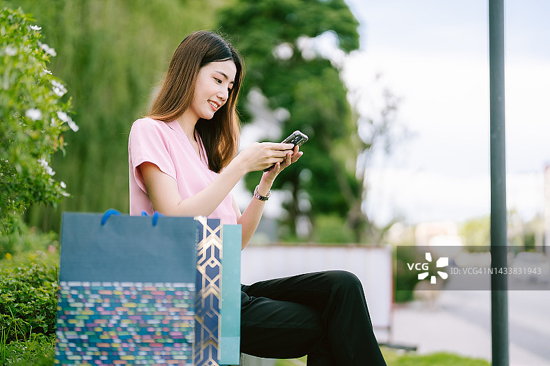 年轻的亚洲妇女拿着购物袋在商场前快乐地坐在电话在早上图片素材