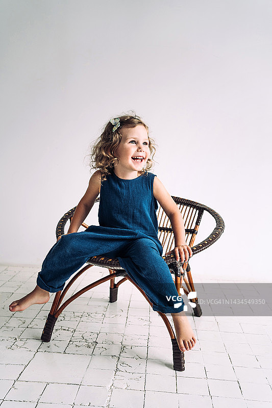金发卷发的小女孩坐在靠白墙的柳条椅上笑。无忧无虑的童年观念图片素材