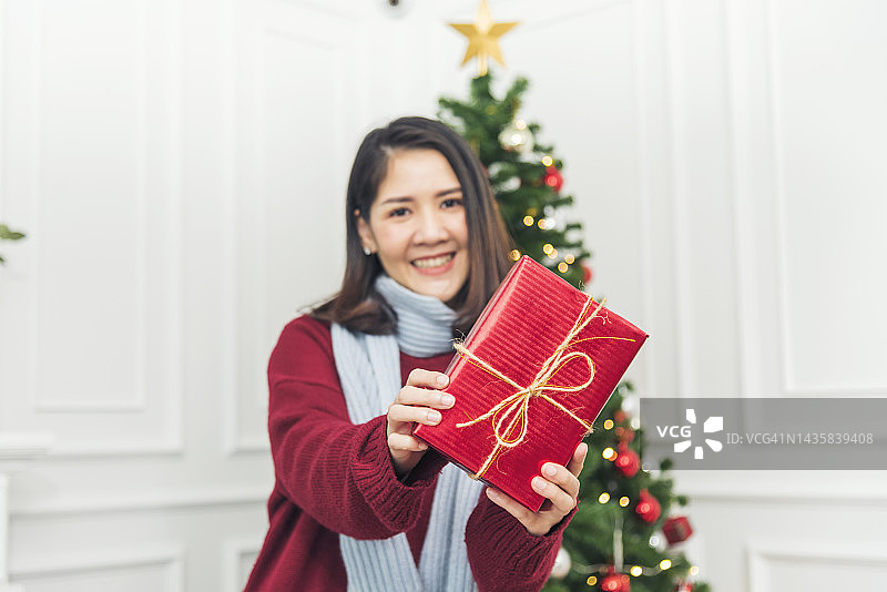圣诞节前夕，一名女子惊讶地收到了礼物盒。亚洲妇女拿着圣诞礼品盒在圣诞树下与微笑的脸。妇女用手包裹礼物。圣诞快乐新年快乐的概念图片素材