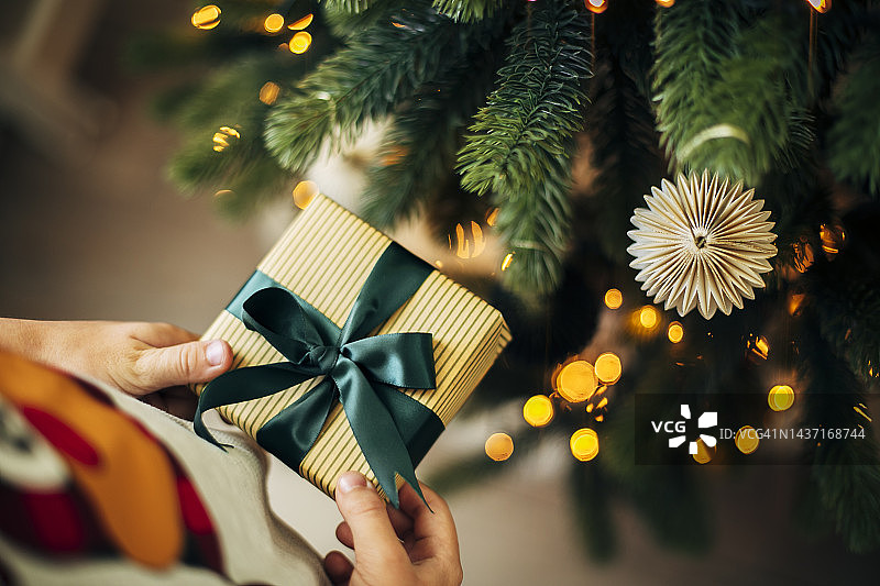 在圣诞树旁，孩子的手拿着包装好的礼品盒图片素材
